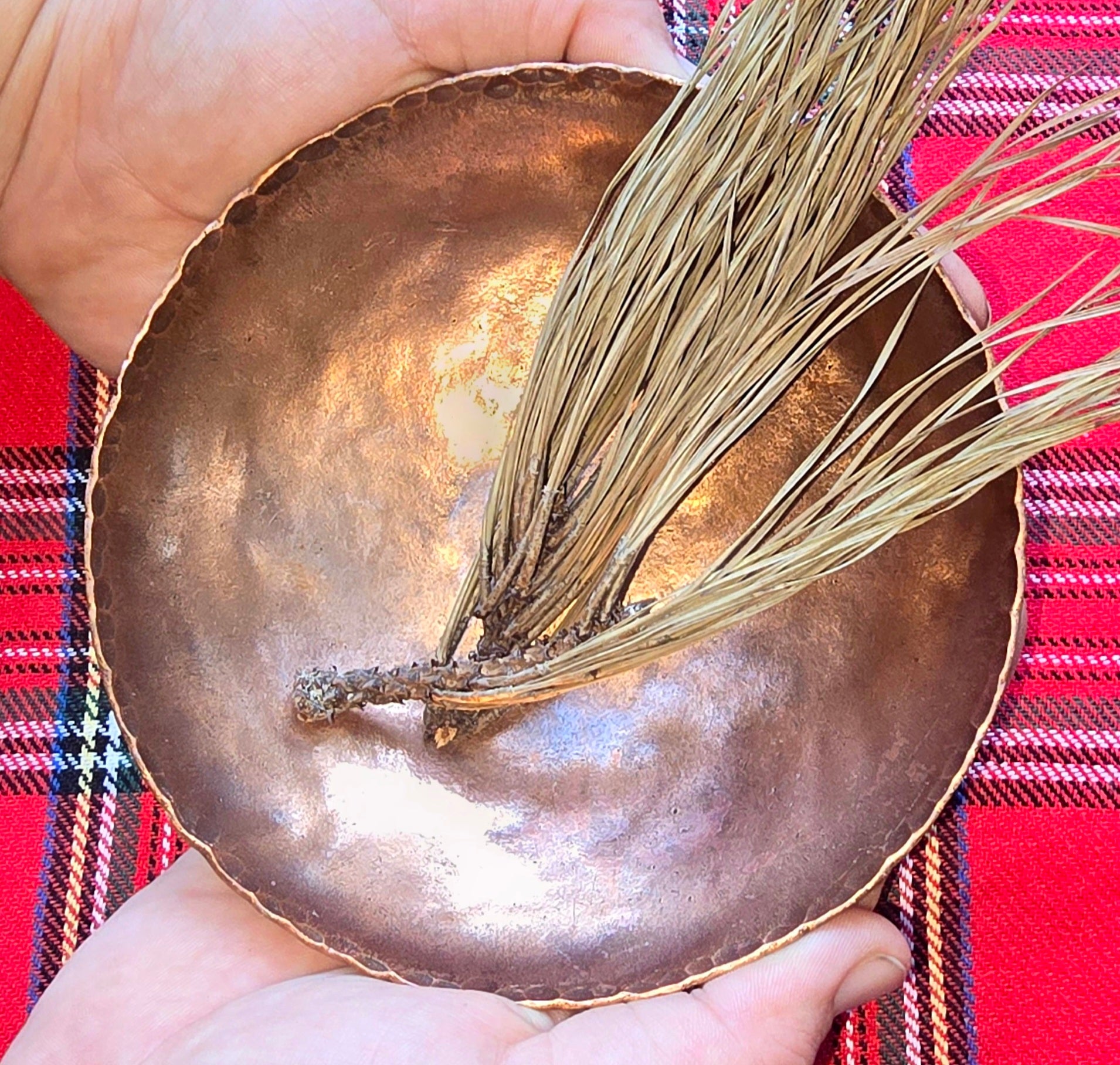 Handcrafted copper bowl in hands with pine needles on red plaid. Artisan rustic home decor.