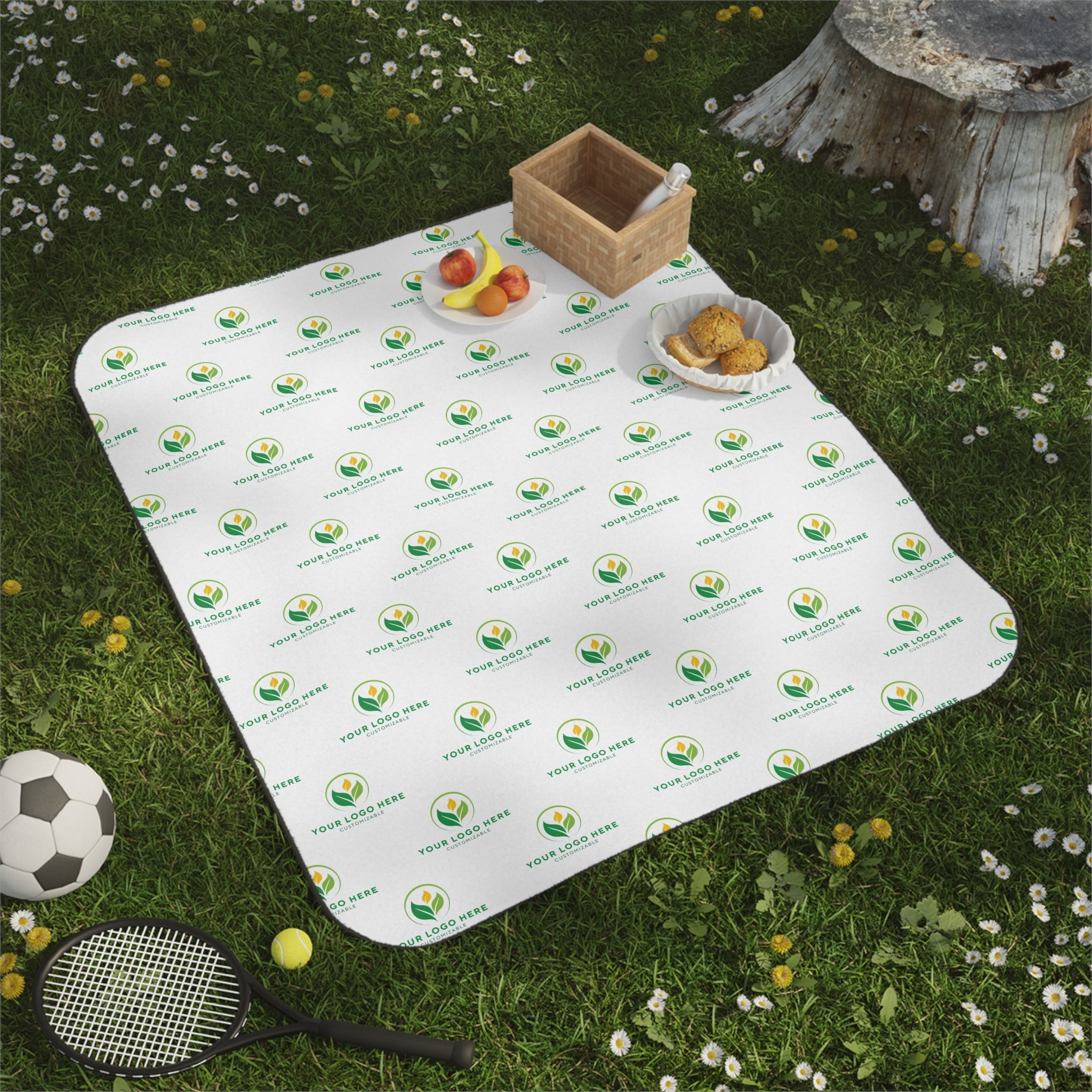 Customizable picnic blanket with company logo on grass with basket, fruits, tennis racket and soccer ball. Outdoor brand promotion.