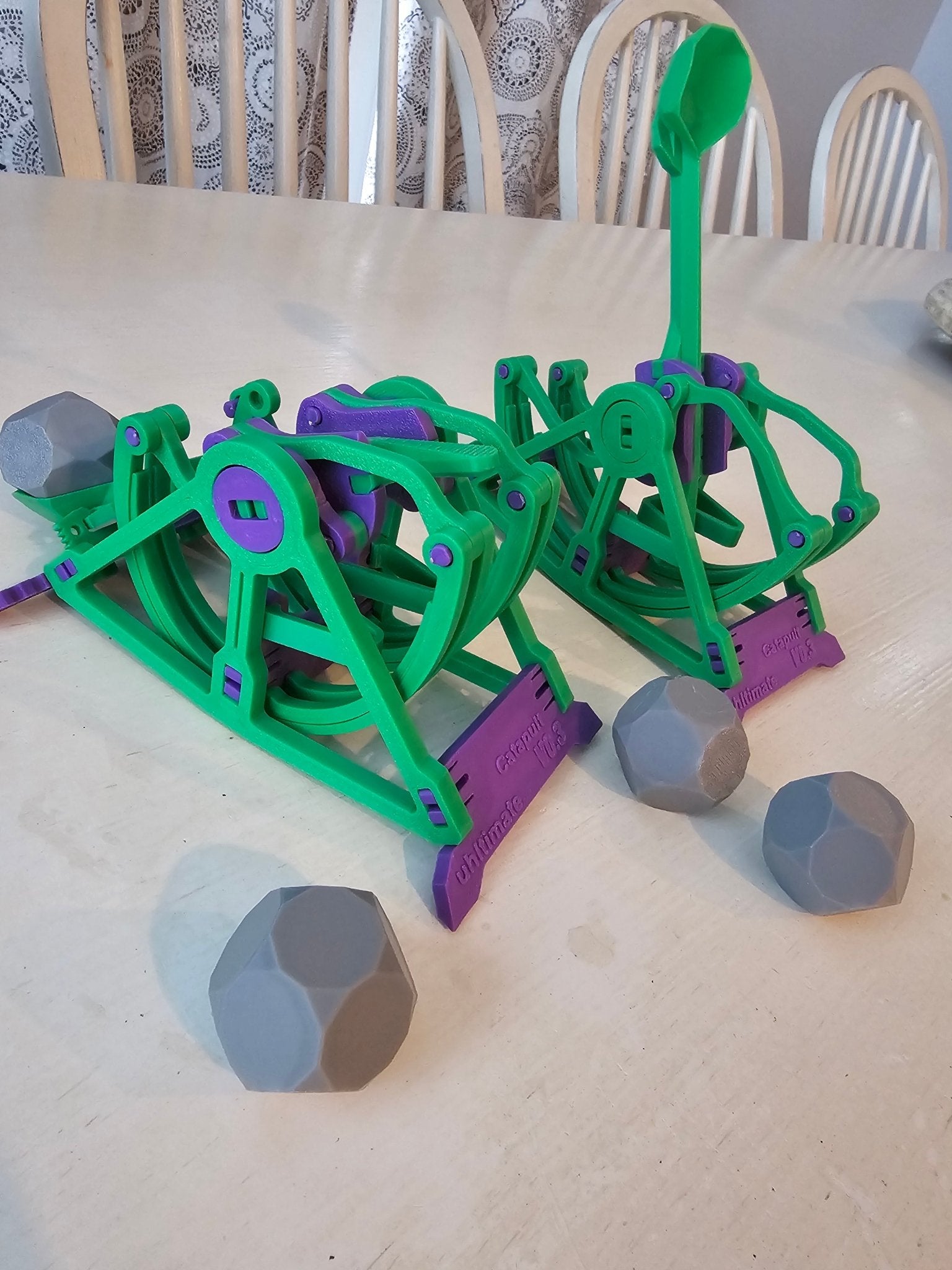 3D-printed catapult launcher toy with green and purple parts and gray boulder projectiles on a table.