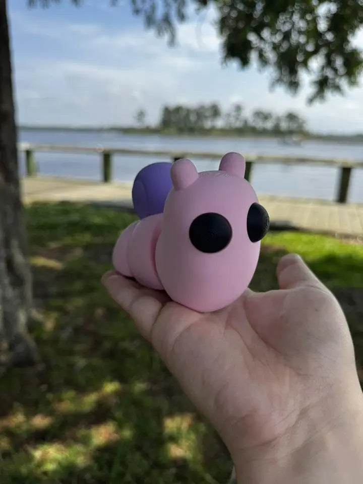 Pink and purple 3D printed snail fidget toy held in hand with lake and trees in background.