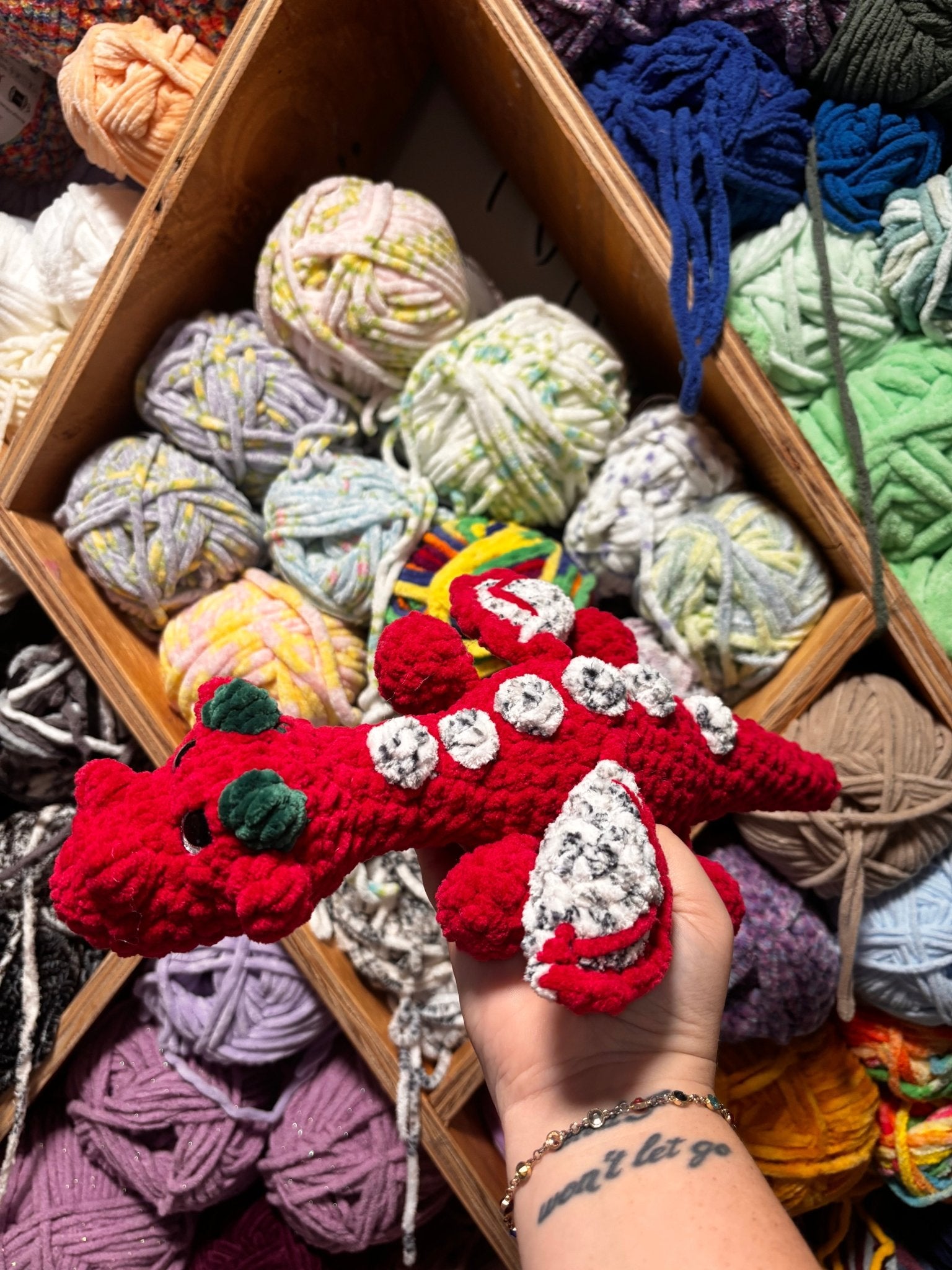 Handmade crochet red dragon plush toy in front of colorful yarn. 'Fruity the Dragon' from Main Street Collective.