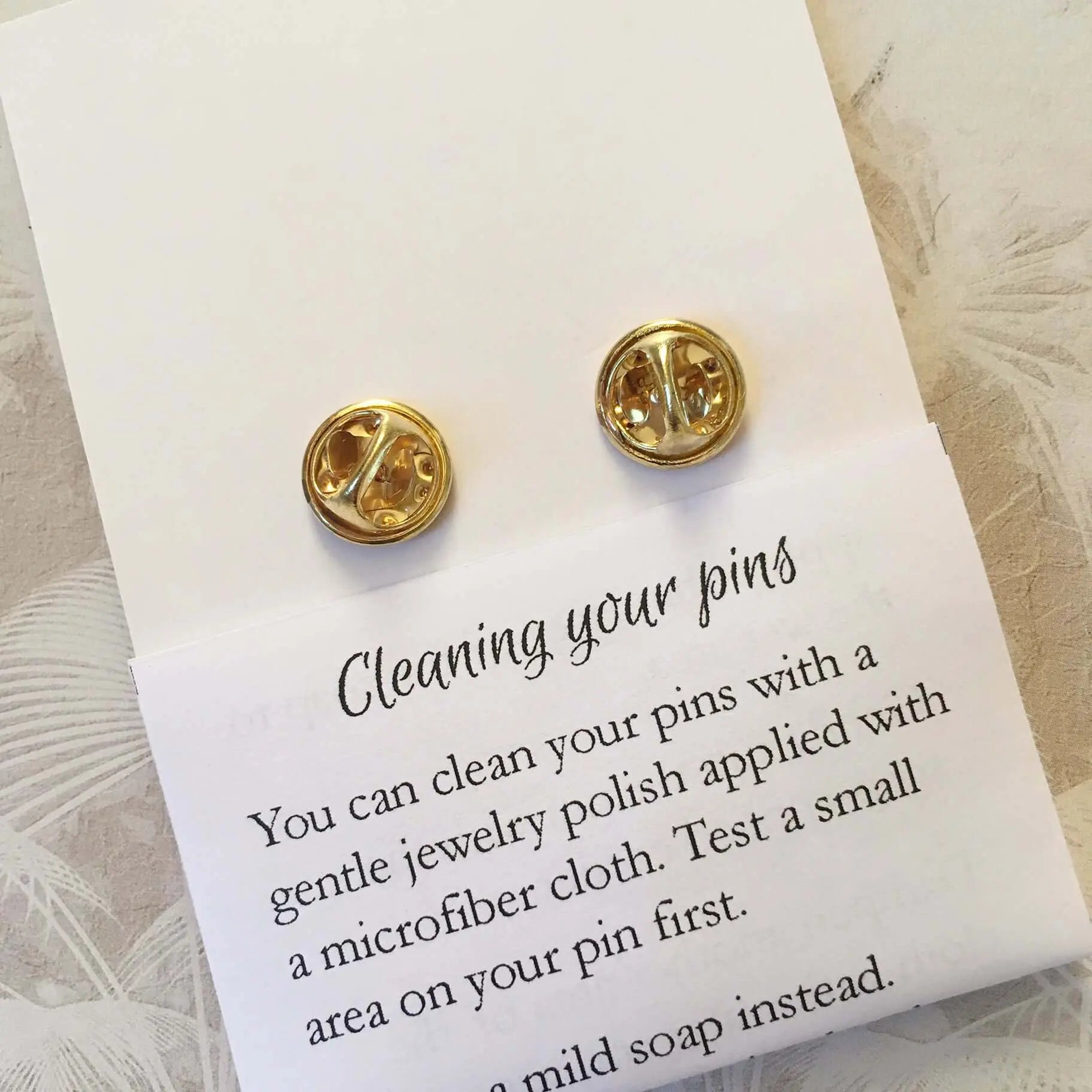 Enamel Pin Backs: Close-up of gold pin backs with care instructions - "Cleaning your pins" guide included.