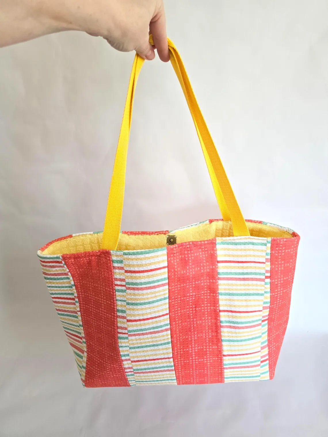 Striped terry cloth beach tote with coral, yellow, and blue stripes, and yellow straps being held in front of white background