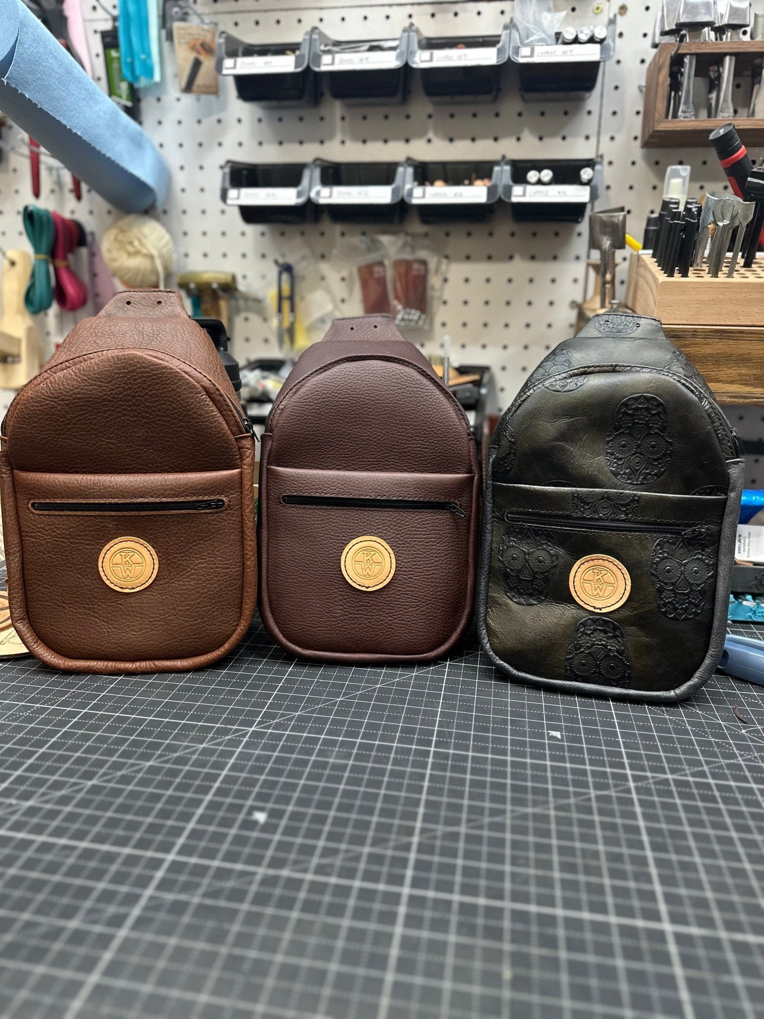 Three handcrafted leather sling bags in brown, dark brown, and skull-embossed black on a black cutting mat.
