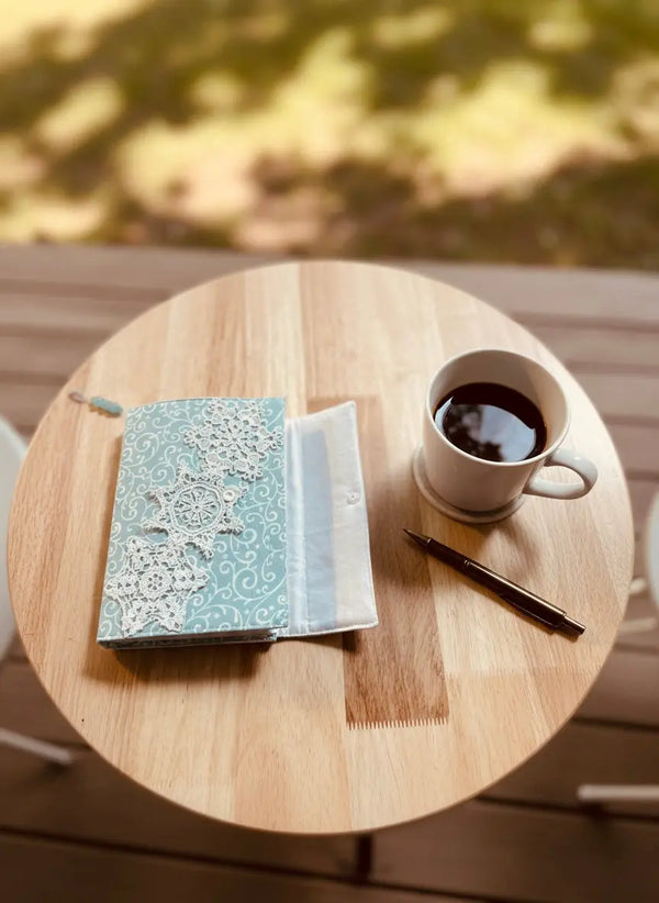 Journal with lace cover, pen, and coffee cup on a wooden table.