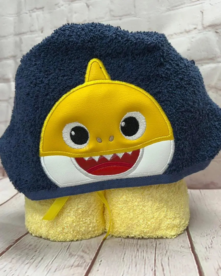 Shark hooded towel for kids: A blue and yellow towel featuring a shark face and a hood, displayed on a wooden background.