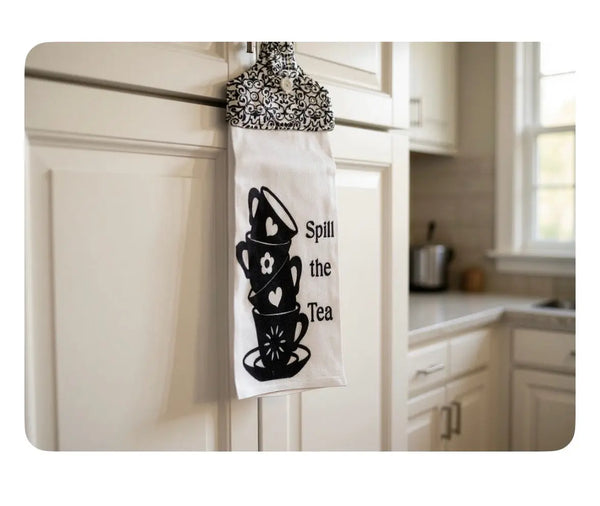 Tea towel hanging on cabinet with stack of teacups and "Spill the Tea" text in a kitchen.